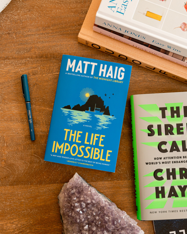 An overhead shot shows a bunch of books on a coffee table including Matt Haig's "The Life Impossible" and Chris Hayes' "The Sirens' Call"