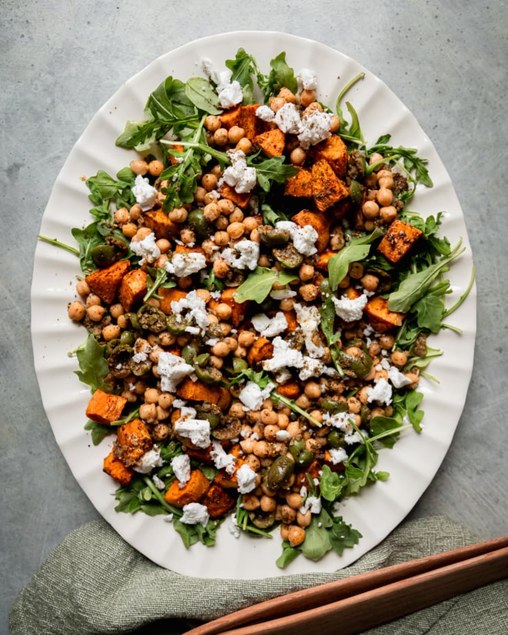 An overhead shot shows a roasted sweet potato salad with baby arugula, vegan "feta" cheese, and a warm olive and chickpea "dressing. The salad is on a white platter and wooden tongs as well as a napkin are seen to the side.