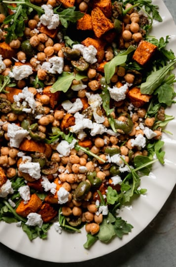 An up close, overhead shot shows a roasted sweet potato salad with baby arugula, vegan "feta" cheese, and a warm olive and chickpea "dressing. The salad is on a white platter.