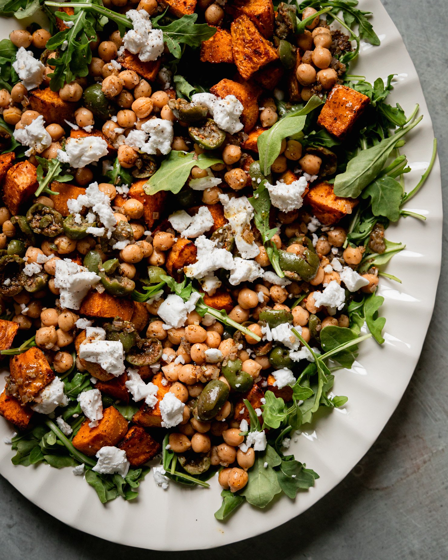 An up close, overhead shot shows a roasted sweet potato salad with baby arugula, vegan "feta" cheese, and a warm olive and chickpea "dressing. The salad is on a white platter.