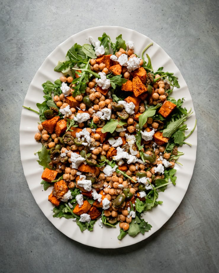 An overhead shot shows a roasted sweet potato salad with baby arugula, vegan "feta" cheese, and a warm olive and chickpea "dressing. The salad is on a white platter against a mottled background.