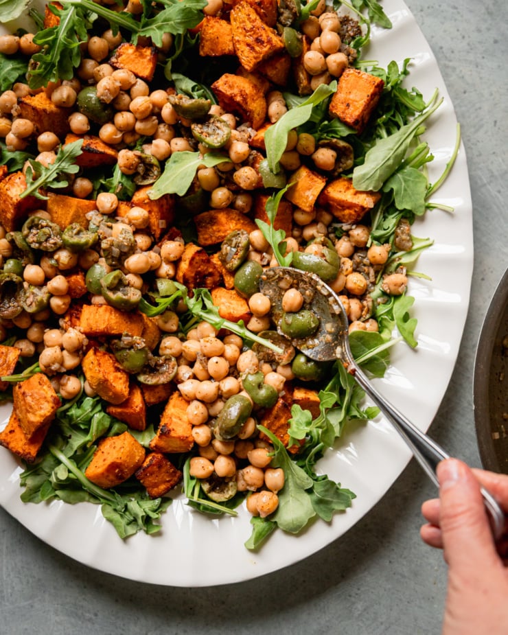 An overhead shot shows a hand spooning a chickpea and olive mixture over arugula and roasted sweet potatoes.