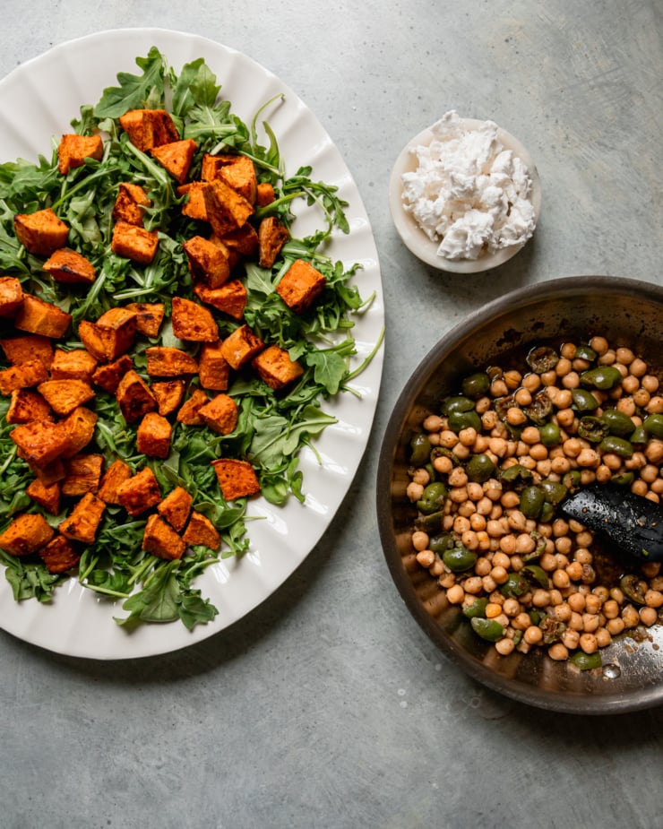 An overhead shot shows: a platter with arugula and roasted sweet potatoes, a small bowl of crumbled vegan "feta" cheese, and a sauté pan with an oily mixture of chopped green olives, chickpeas, shallots, and za'atar.