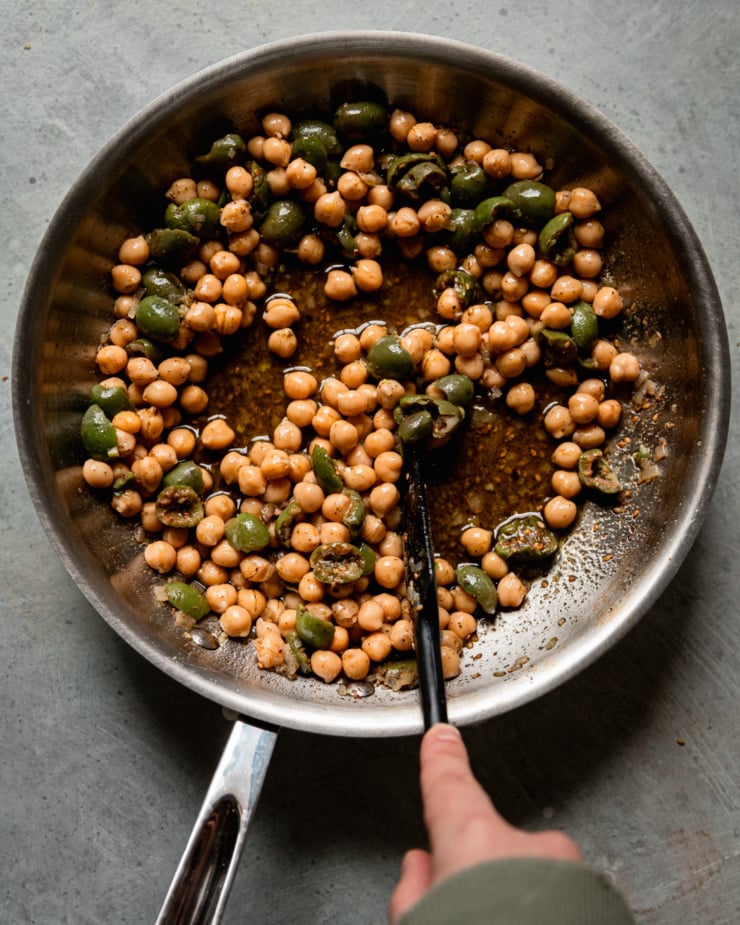 An overhead shot shows a hand stirring a mixture of green olives, chickpeas, shallots, za'atar, and olive oil with a spatula in a sauté pan.