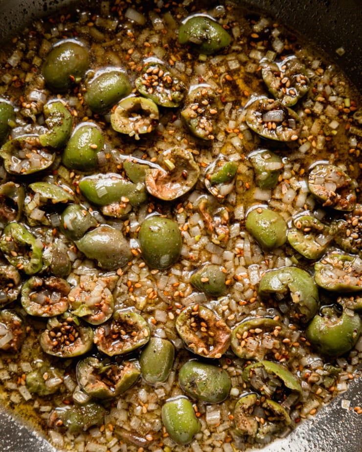 An up close, overhead shot shows a sauté pan containing a mixture of olive oil, minced shallots, chopped green olives, and za'atar.