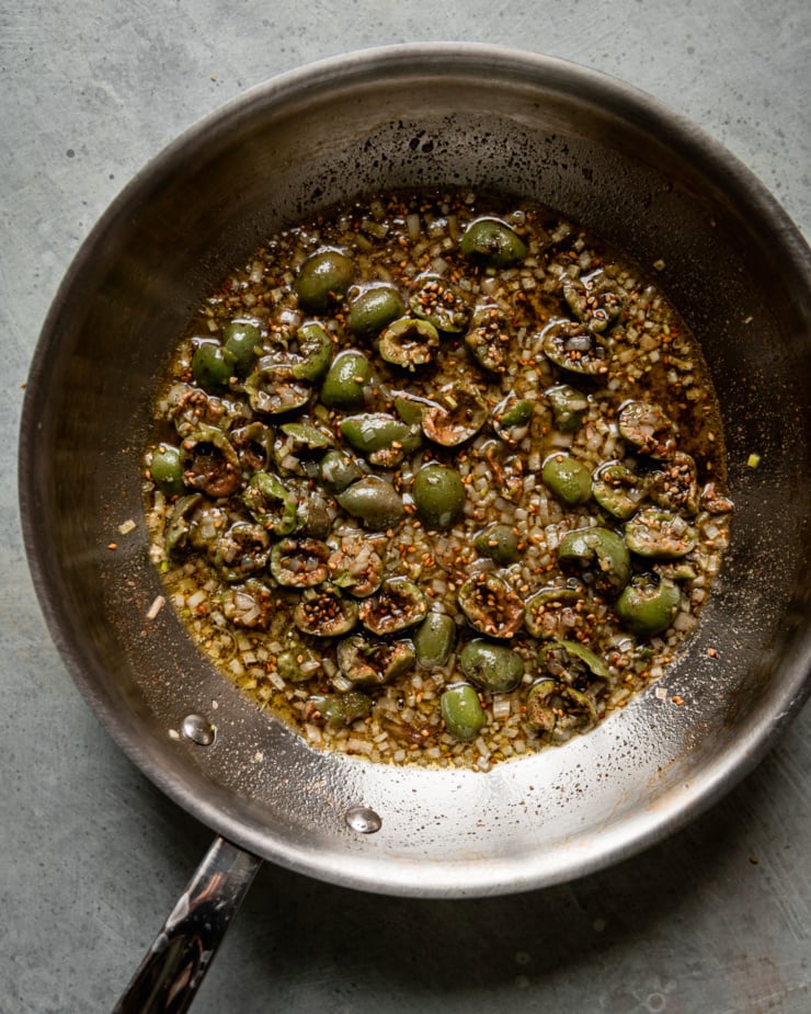 An overhead shot shows a sauté pan containing a mixture of olive oil, minced shallots, chopped green olives, and za'atar.
