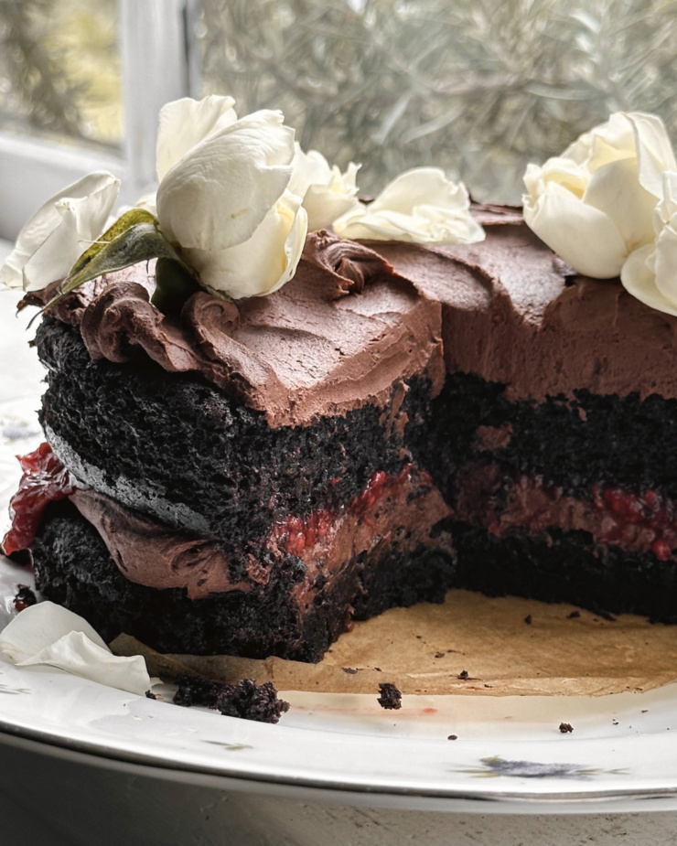A head-on shot shows a vegan chocolate cake with black tea ganache and raspberry rose compote. The cake has been cut into, revealing the beautiful layers within. White flowers adorn the top.
