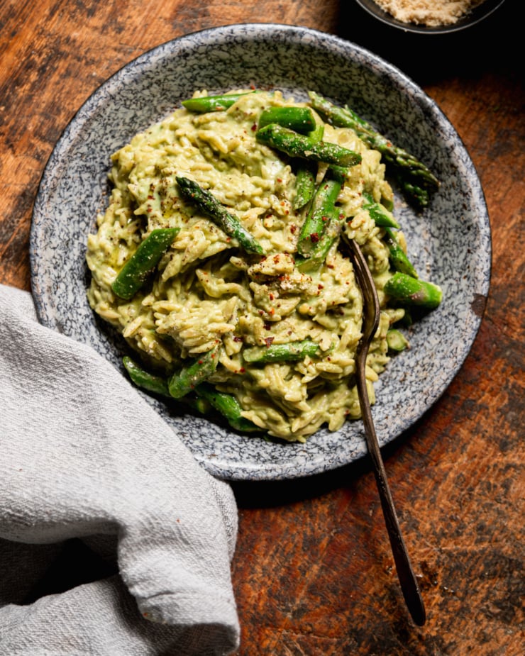 An overhead shot shows a serving of spring green orzo risotto topped with asparagus. A fork is sticking out of the serving in a shallow enamelware bowl.