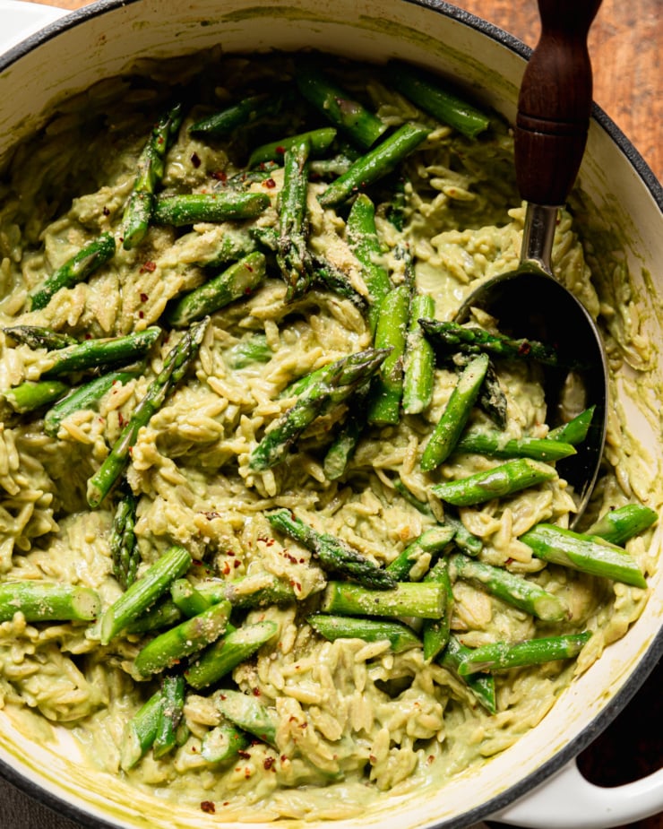 An up close, overhead shot shows a pot of spring green orzo risotto with sautéed asparagus. A serving spoon is sticking out of the pot.