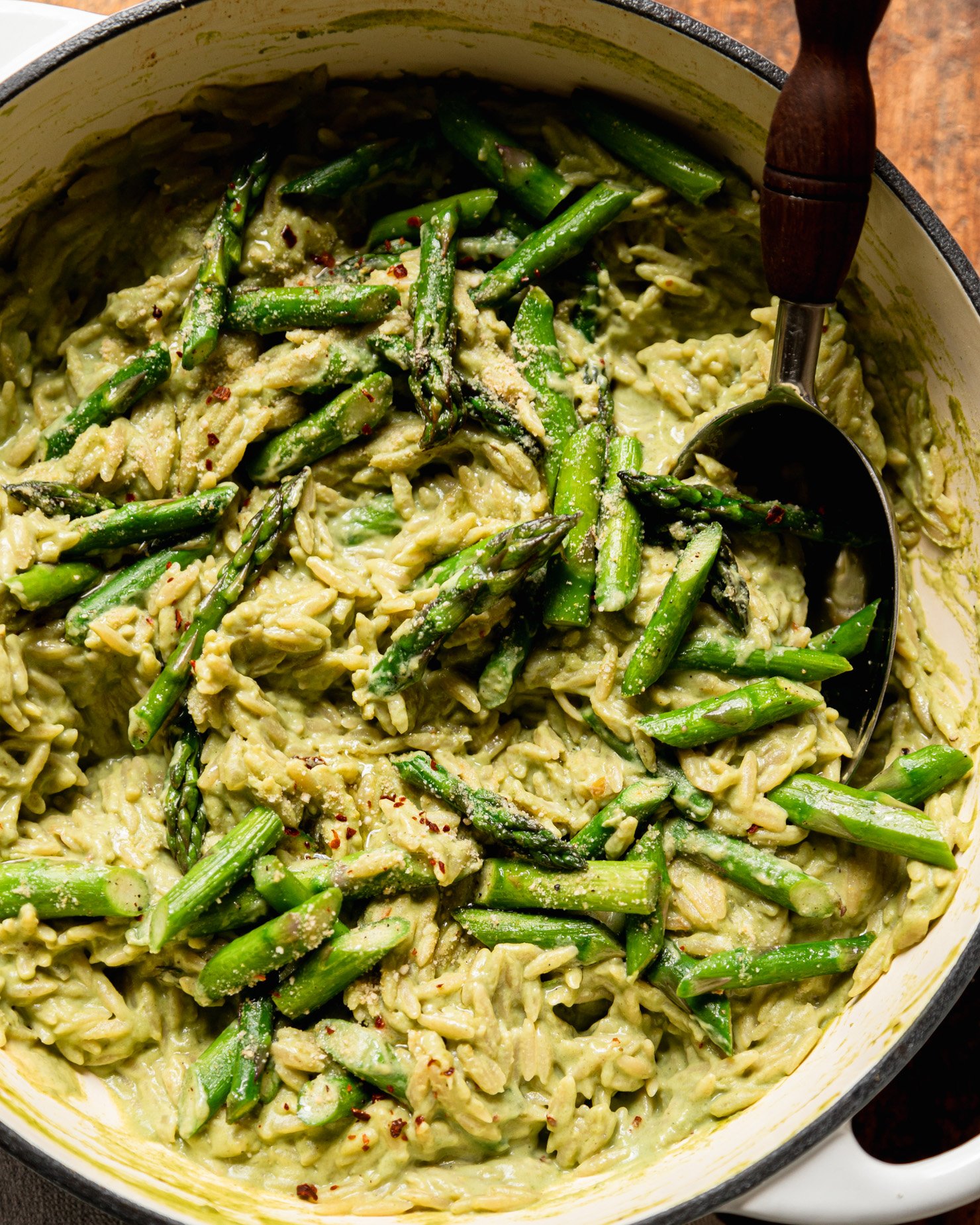 An up close, overhead shot shows a pot of spring green orzo risotto with sautéed asparagus. A serving spoon is sticking out of the pot.