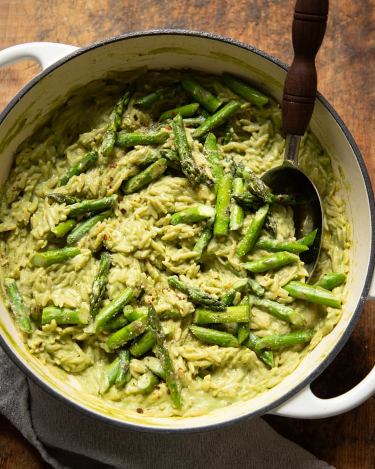An overhead shot shows a pot of spring green orzo risotto with sautéed asparagus. A serving spoon is sticking out of the pot.