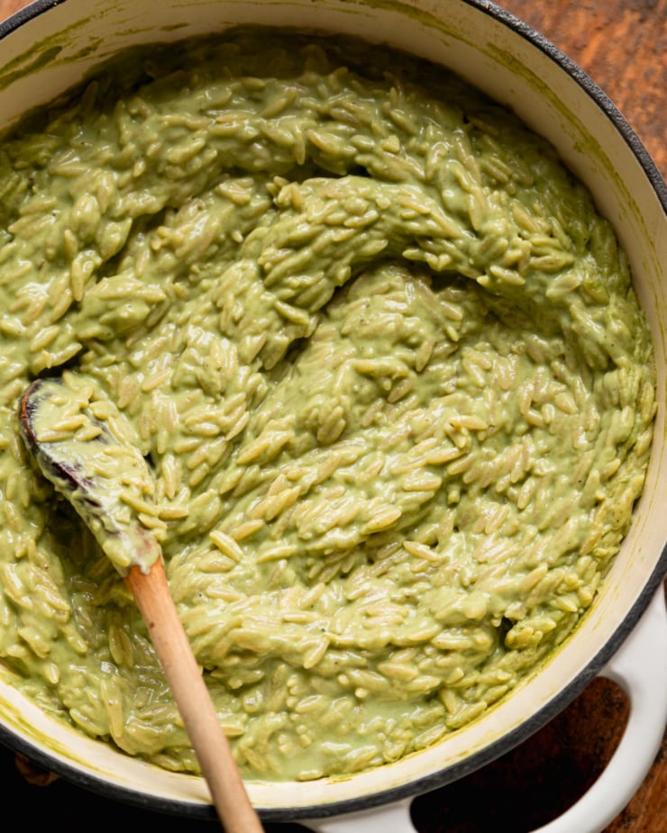 An overhead shot shows a pot of creamy green orzo risotto. A wooden spoon sticks out of the pot.