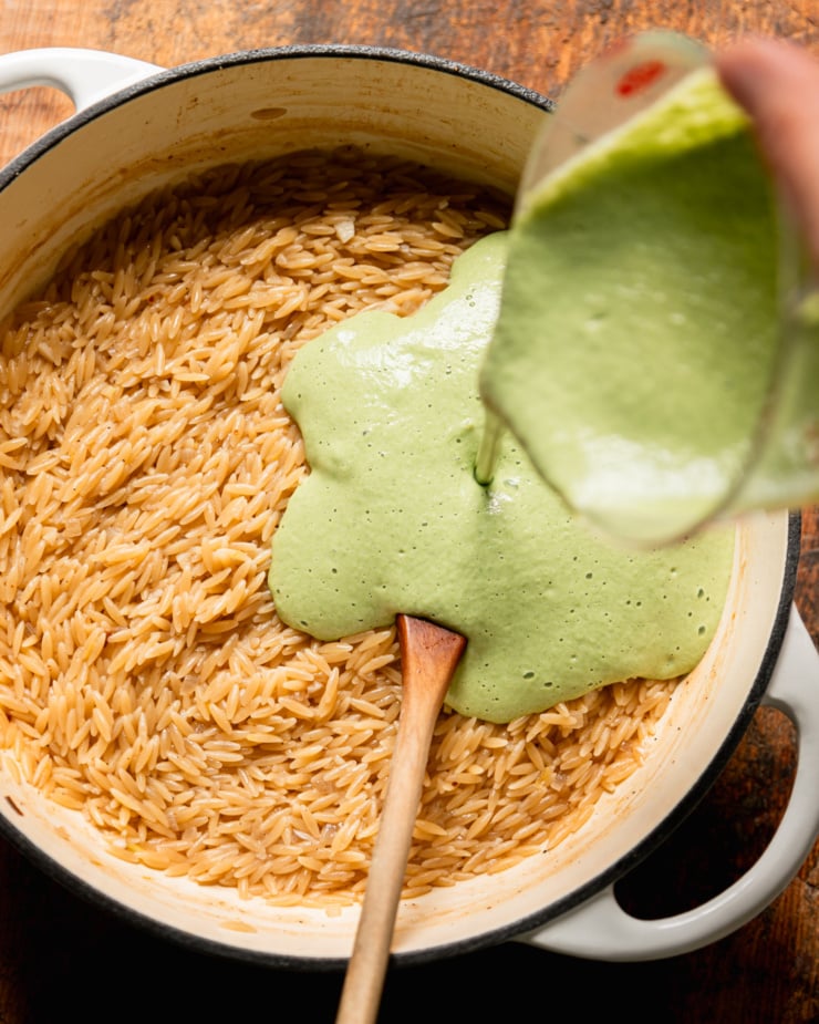 An overhead shot shows a hand pouring a green creamy mixture into a pot of cooked orzo.