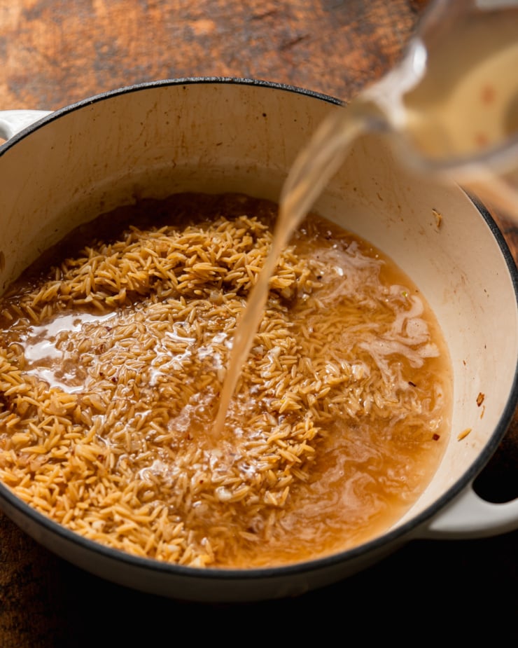 A 3/4 angle shot shows vegetable stock being poured into a pot with sautéed shallots and orzo.