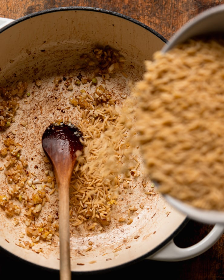 An overhead shot shows orzo pasta being poured into a pot with sautéed shallots and spices.