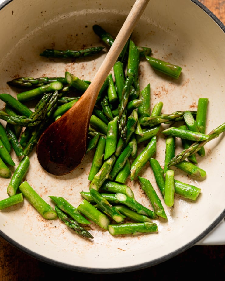 An overhead shot shows sautéed chopped asparagus in a braiser pot with a wooden spoon.