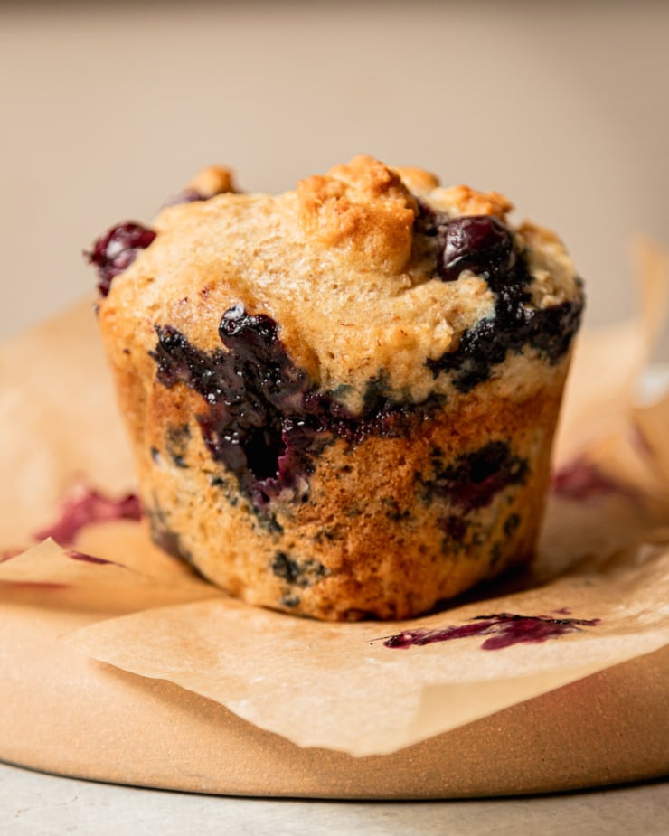 An up close, head-on shot shows a vegan blueberry muffin. The muffin's liner has been peeled off and you can see a big oozy pocket of blueberry on the side.