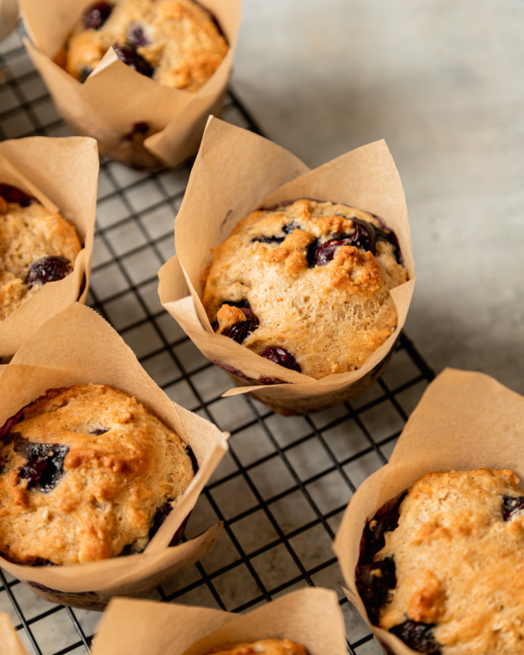 A 3/4 angle shot shows perfectly puffed up vegan blueberry muffins on a wire cooling rack. The muffins are wrapped in parchment paper liners.