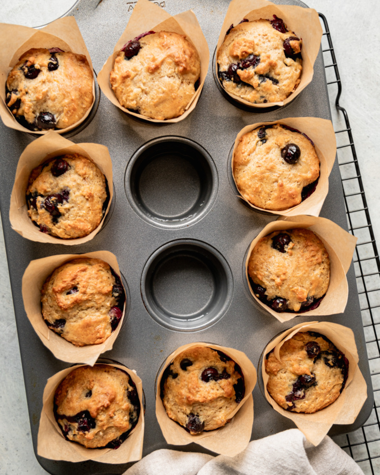 An overhead shot shows freshly baked vegan blueberry muffins in a muffin tin. The center cups of the muffin tin are filled with hot water. The tin is resting on a wire cooling rack.