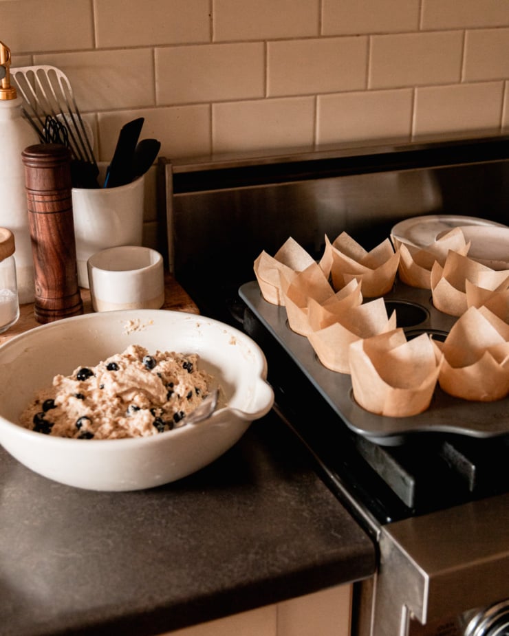 A head-on shot shows a bowl of batter on a kitchen counter next to a muffin tin with homemade parchment liners inside each cavity.