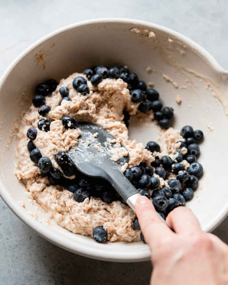 An overhead shot shows a hand using a spatula to fold fresh blueberries into a batter in a large bowl.