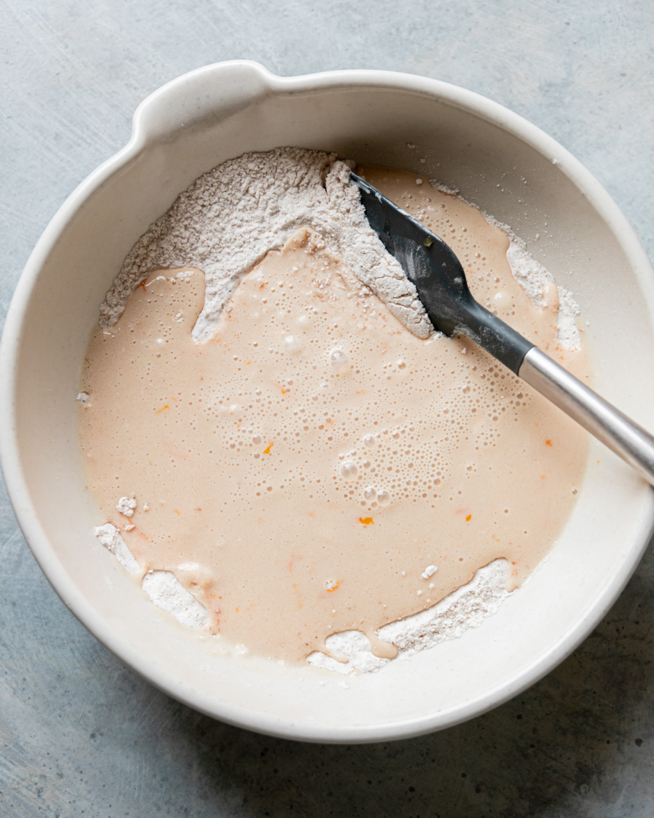 An overhead shot shows a orange zest-flecked wet mixture that's been poured on top of a flour mixture in a large bowl. A rubber spatula is sticking out of the bowl.