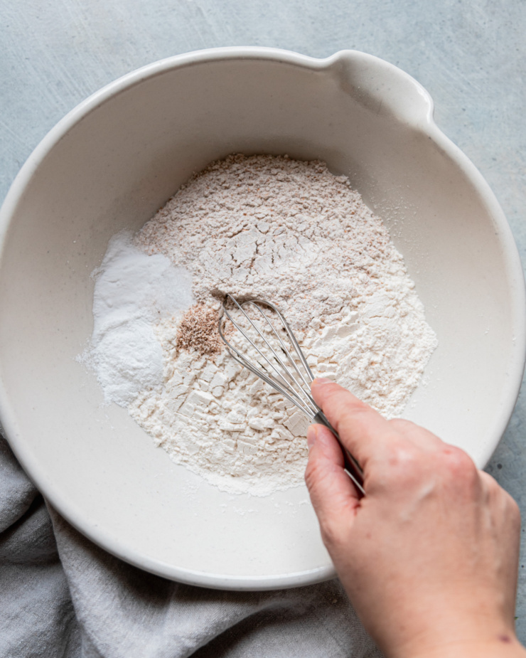 An overhead shot shows a hand using a whisk to combine dry ingredients in a large bowl with a pouring spout.