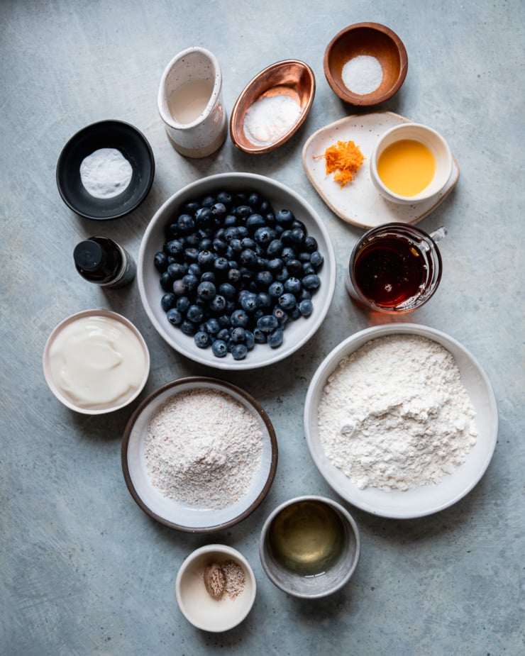 An overhead shot shows ingredients for vegan blueberry muffins, all prepped and measured in small bowls. There's baking powder, baking soda, orange zest & juice, maple syrup, all purpose flour, sunflower oil, nutmeg, whole wheat flour, vegan yogurt, vanilla extract, fresh blueberries, salt, and soy milk.