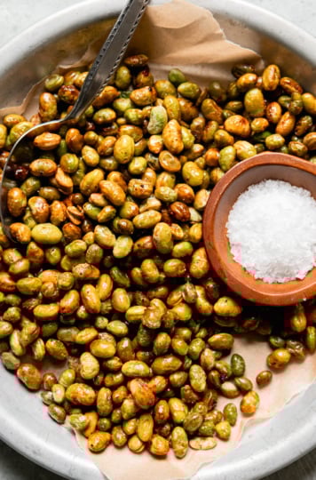 An overhead shot shows a shallow dish lined with paper, filled with air fryer crispy edamame. The edamame are lightly browned and a small bowl of flaky salt is seen to the side.