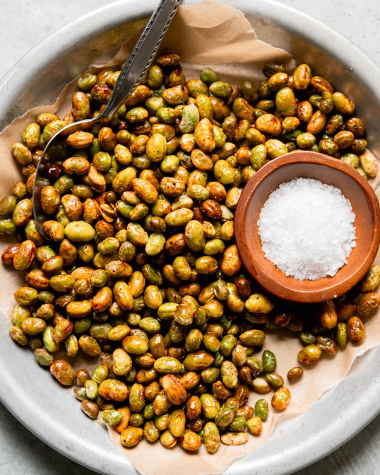 An overhead shot shows a shallow dish lined with paper, filled with air fryer crispy edamame. The edamame are lightly browned and a small bowl of flaky salt is seen to the side.