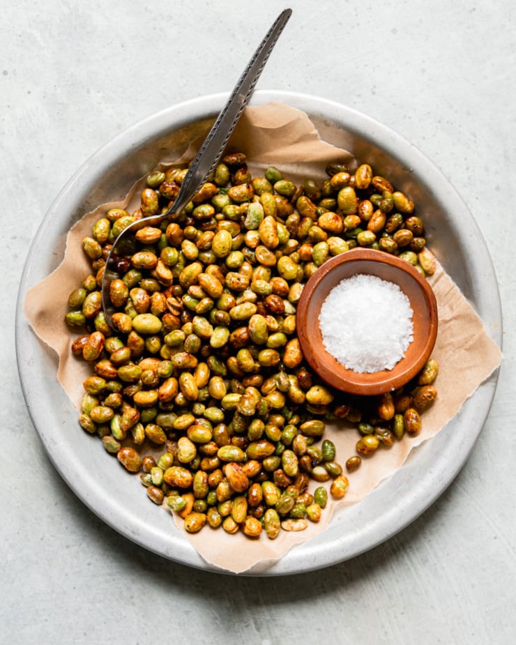 An overhead shot shows a shallow dish lined with paper, filled with air fryer crispy edamame. The edamame are lightly browned and a small bowl of flaky salt is seen to the side.