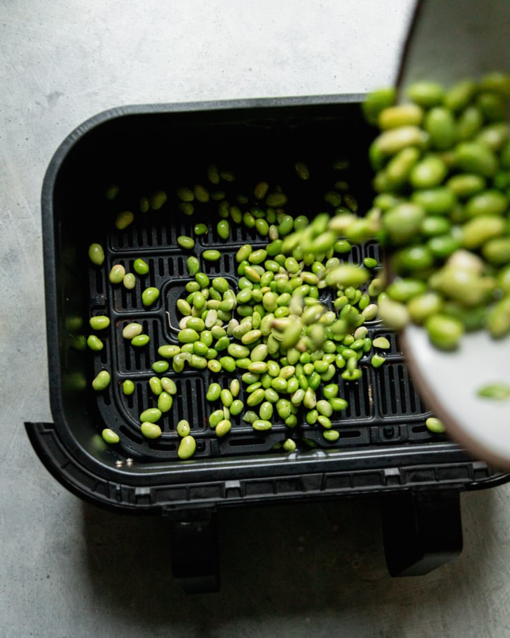 An overhead shot shows edamame being poured into an air fryer basket from above.