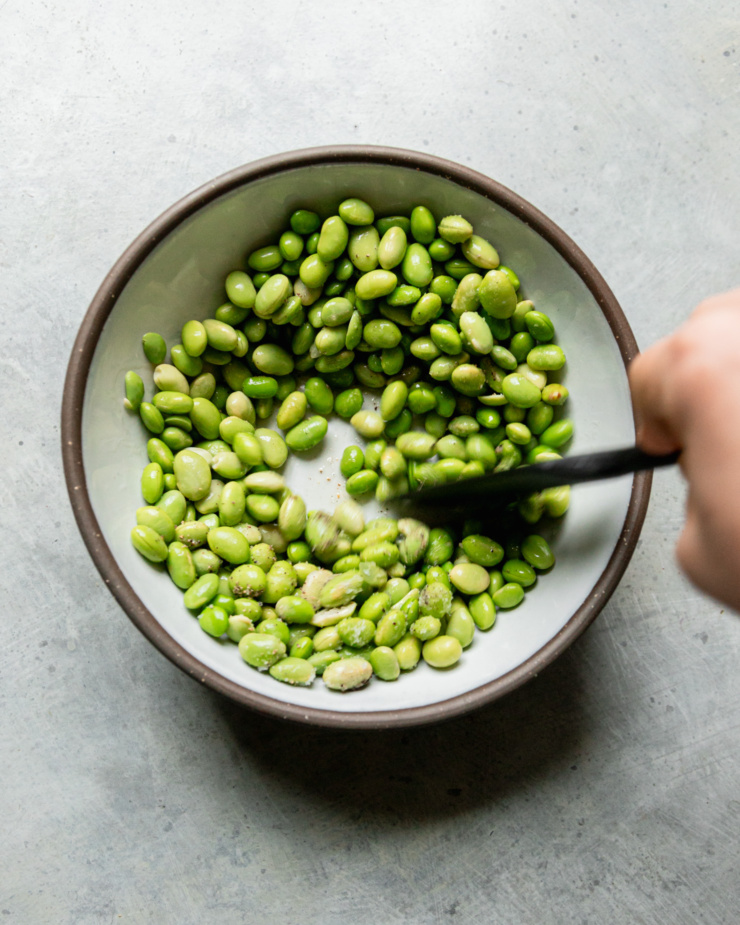 An overhead shot shows a hand using a spatula to stir some shelled edamame with oil, salt, and pepper.