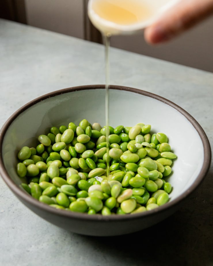 A head-on shot shows oil being poured over a bowl of shelled edamame.