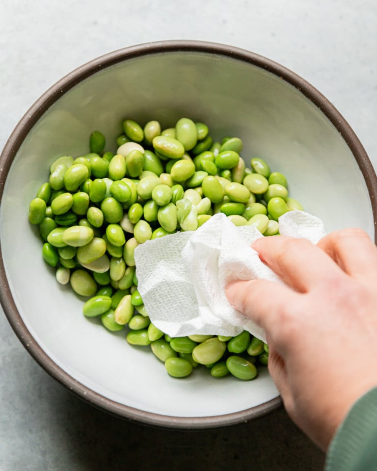 An overhead shot shows a hand using a paper towel to blot dry some thawed edamame.