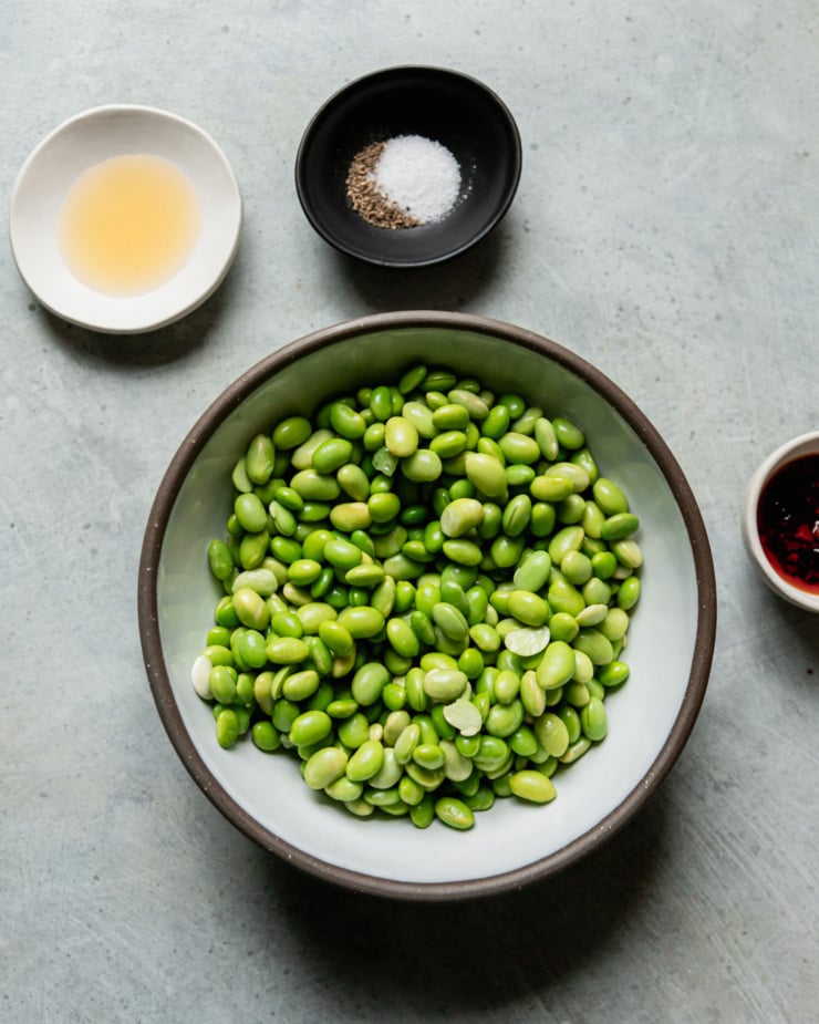 An overhead shot shows a bowl of thawed frozen shelled edamame, a bowl of avocado oil, a bowl with salt and pepper, and a small bowl of chili crisp to the side.