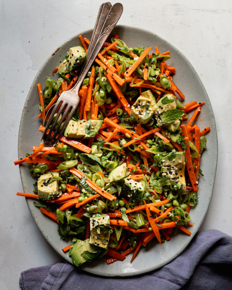 An overhead shot shows a crunchy shredded snap peas and carrots salad with chunks of avocado on top. The salad is dressed with a sesame, lime and garlic dressing. Chopped cilantro, Thai basil, and toasted sesame seeds fleck the salad throughout. The salad is on a platter with a fork and spoon for tongs presented on the side of the plate.