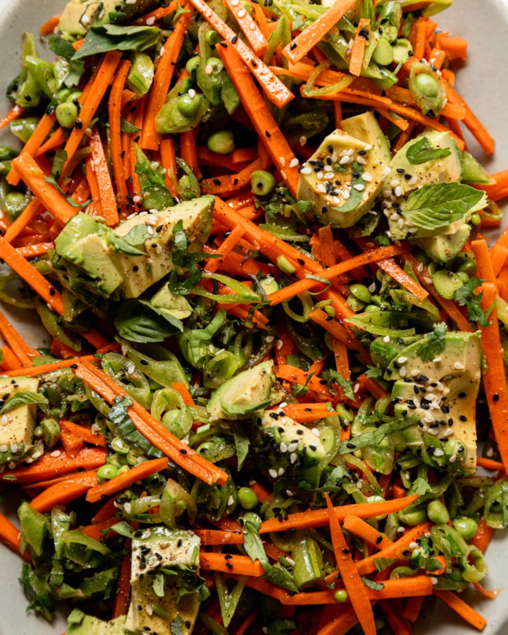 An up close, overhead shot shows a crunchy shredded snap peas and carrots salad with chunks of avocado on top. The salad is dressed with a sesame, lime and garlic dressing. Chopped cilantro, Thai basil, and toasted sesame seeds fleck the salad throughout.