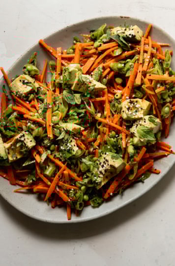An overhead shot shows a crunchy shredded snap peas and carrots salad with chunks of avocado on top. The salad is dressed with a sesame, lime and garlic dressing. Chopped cilantro, Thai basil, and toasted sesame seeds fleck the salad throughout.