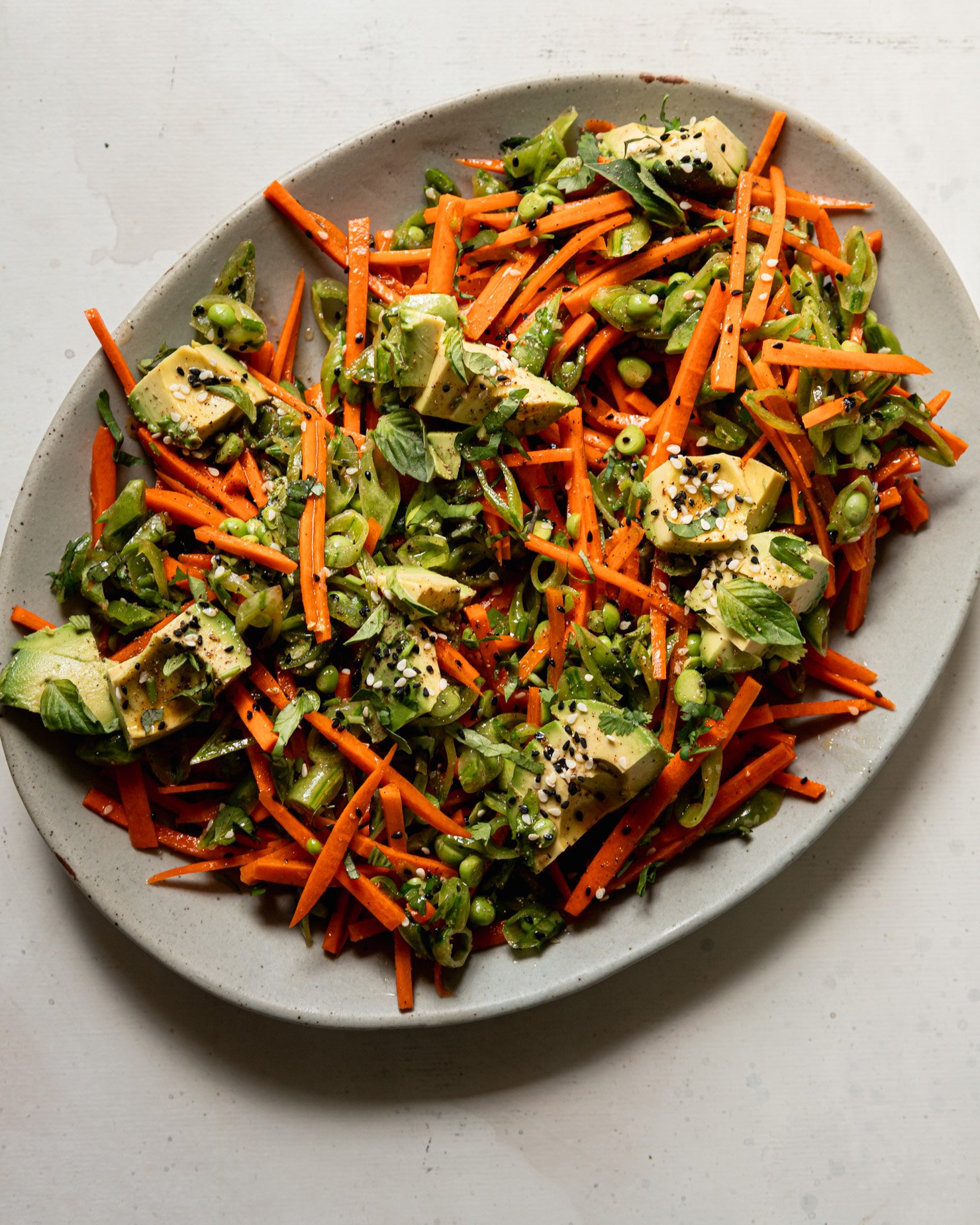 An overhead shot shows a crunchy shredded snap peas and carrots salad with chunks of avocado on top. The salad is dressed with a sesame, lime and garlic dressing. Chopped cilantro, Thai basil, and toasted sesame seeds fleck the salad throughout.