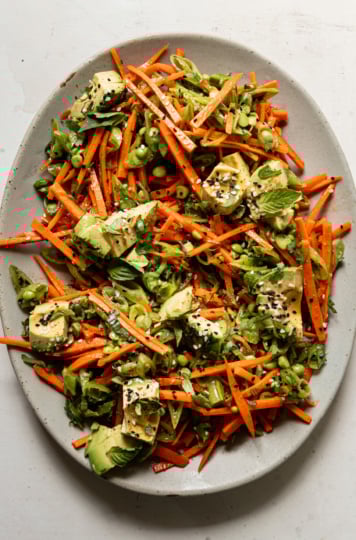 An overhead shot shows a crunchy shredded snap peas and carrots salad with chunks of avocado on top. The salad is dressed with a sesame, lime and garlic dressing. Chopped cilantro, Thai basil, and toasted sesame seeds fleck the salad throughout.