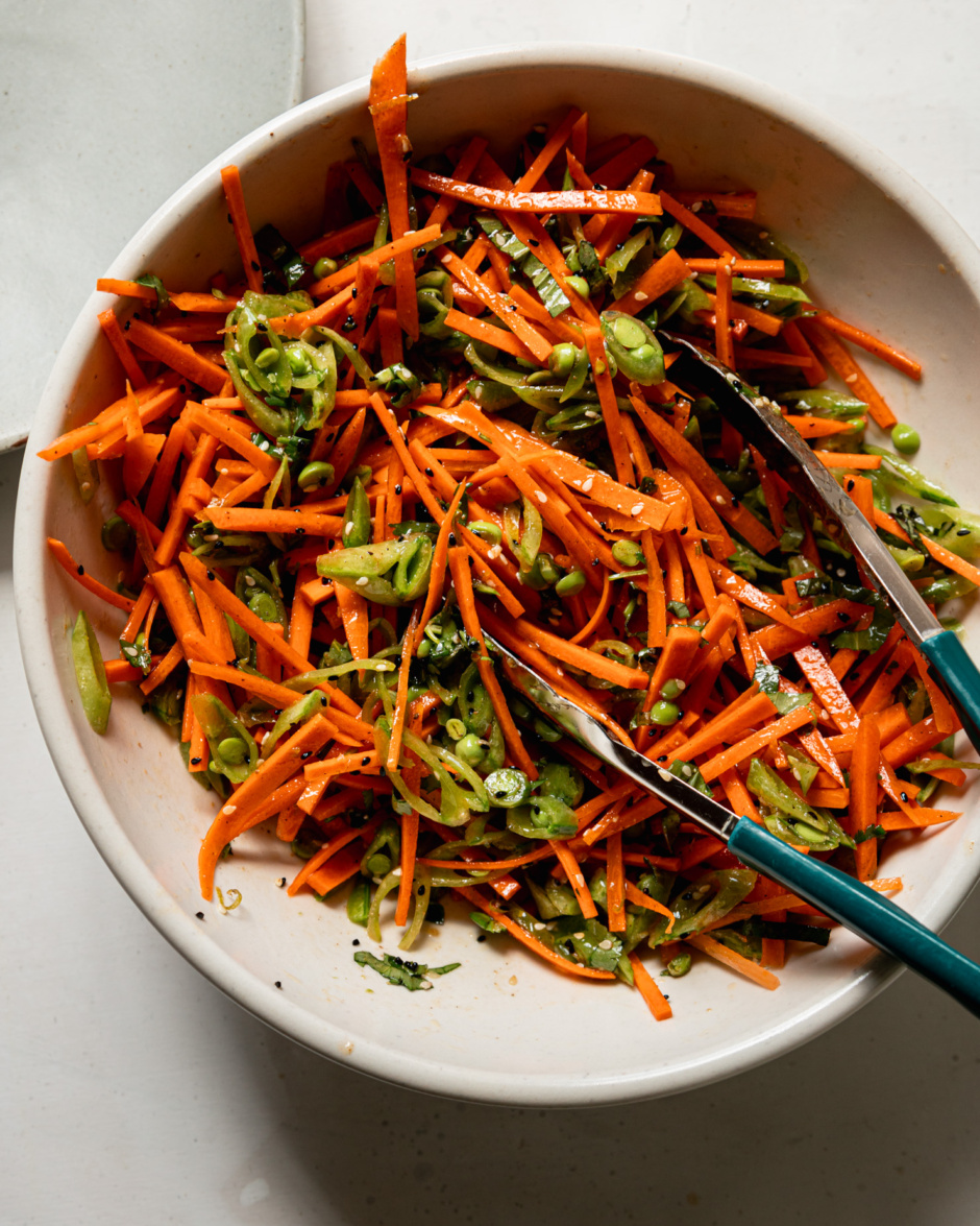 An overhead shot shows a mixing bowl filled with dressed julienned carrots and sliced snap peas. Tongs are sticking out of the bowl.