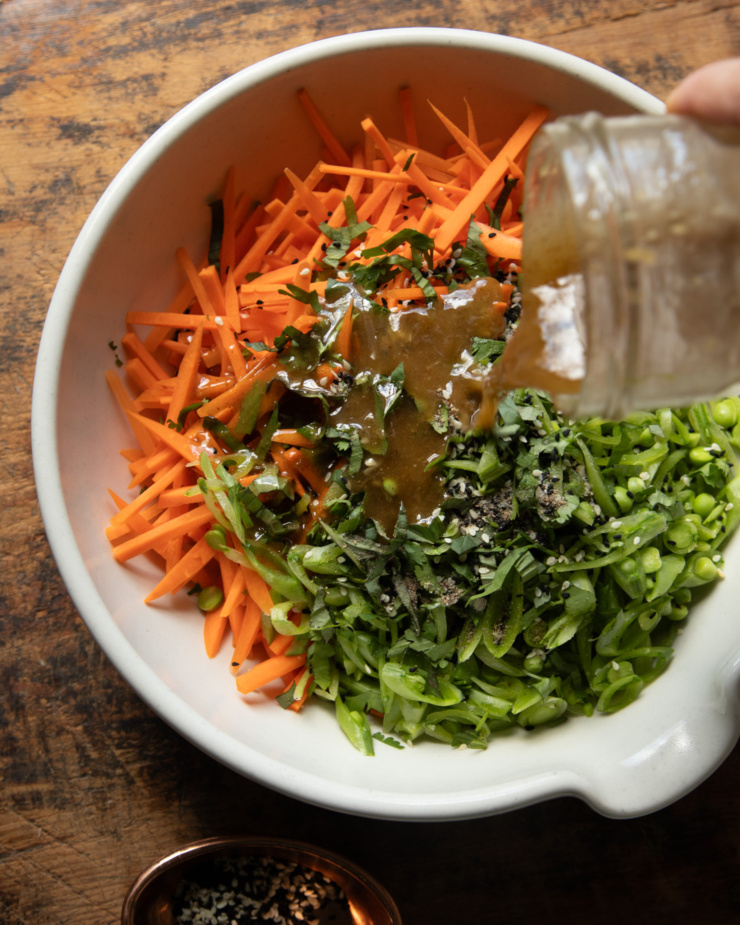 An overhead shot shows a salad dressing being poured over finely sliced vegetables and herbs in a mixing bowl.