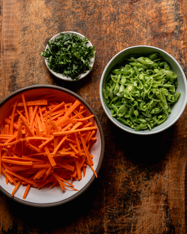 An overhead shot shows: a small bowl of chopped herbs, a medium bowl of sliced raw snap peas, a large bowl of julienned raw carrots.