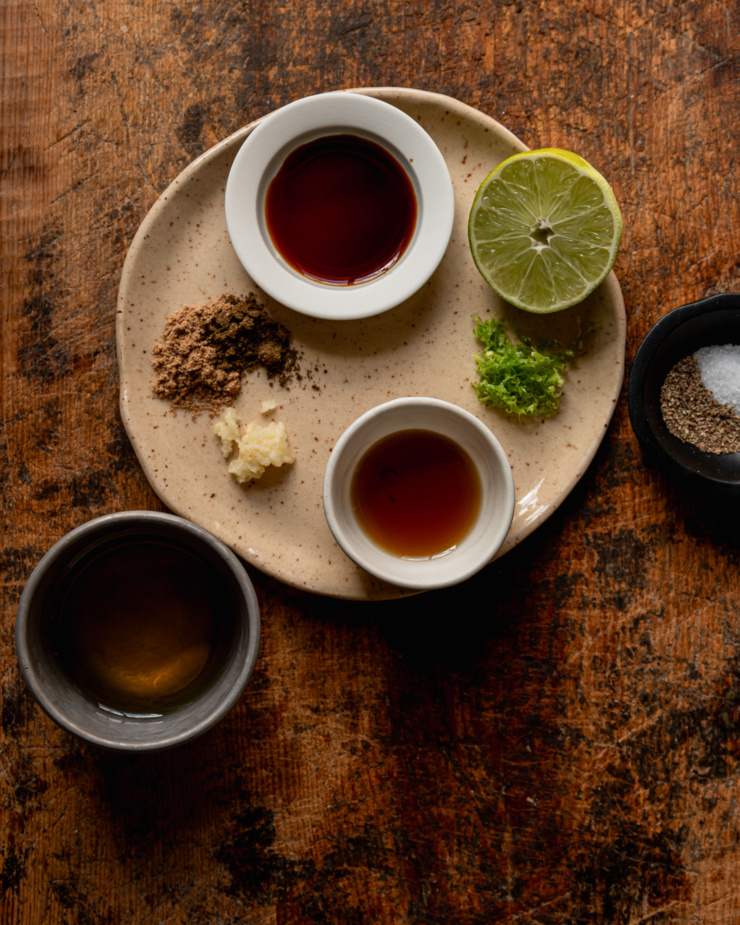 An overhead shot shows ingredients for a sesame lime dressing: Tamari, lime juice, lime zest, sesame oil, garlic, cumin, coriander, salt, pepper, and avocado oil.
