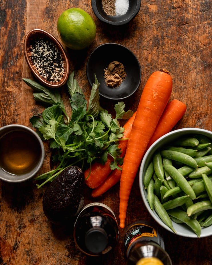 An overhead shot shows ingredients for a salad: carrots, snap peas, sesame oil, Tamari, avocado, cilantro, Thai basil, avocado oil, cumin, coriander, sesame seeds, lime, salt, and pepper. All ingredients are shown on a rough wood cutting board.