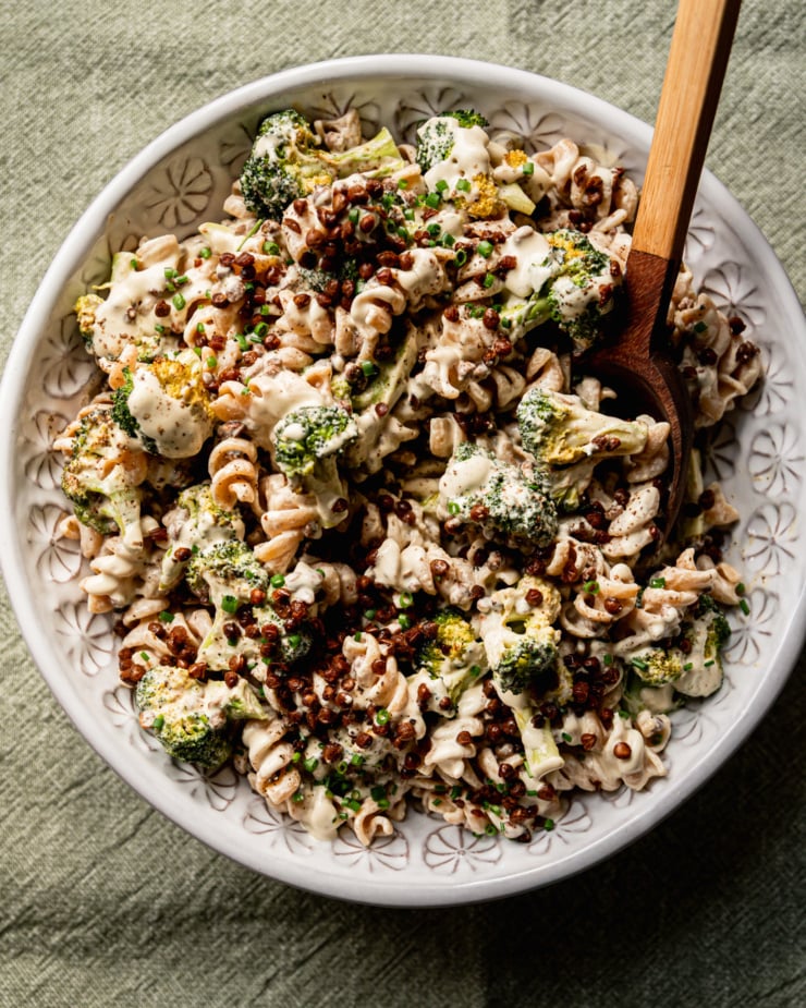 An overhead shot shows a creamy vegan broccoli Caesar pasta salad in a serving bowl. The top of the salad is garnished with chopped chives and roasted smoky lentils, and a wooden serving spoon is dug into the salad.