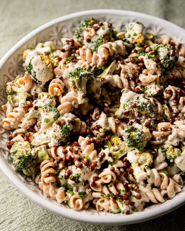 An up close, 3/4 angle shot shows a creamy vegan broccoli Caesar pasta salad in a serving bowl. The top of the salad is garnished with chopped chives. Roasted lentils are also seen on top.