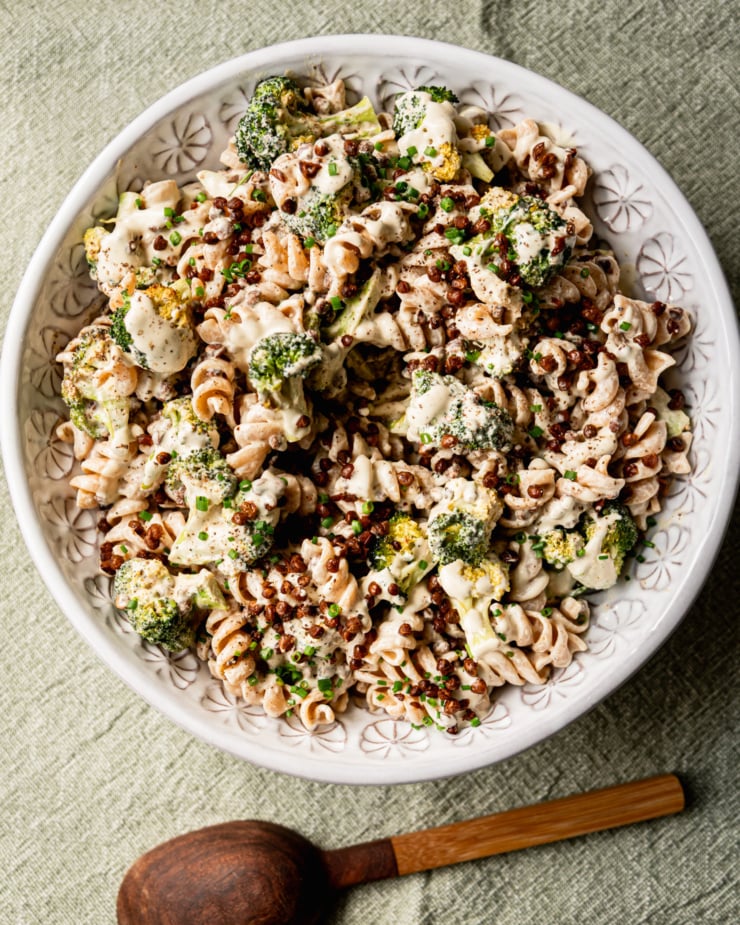 An overhead shot shows a creamy vegan broccoli Caesar pasta salad in a serving bowl. The top of the salad is garnished with chopped chives and a wooden serving spoon is seen to the side.