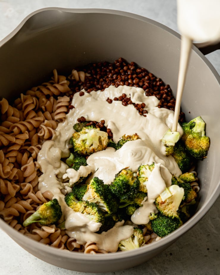 An overhead shot shows vegan Caesar dressing being poured over roasted broccoli florets, lentils, and cooked rotini pasta.